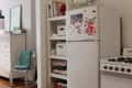 Small kitchen with white fridge, stove, and open shelving. Fridge has magnets, and shelves hold books and baskets.