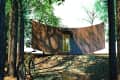 Curved wooden cabin in a forest with a bicycle parked nearby, surrounded by tall trees.