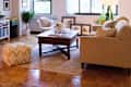 Cozy living room with beige sofa, wooden coffee table, yellow pouf, and large windows. Decor includes plants and framed art.