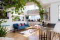 Bright living room with blue sofa, colorful cushions, hanging plants, dartboard, and bar stools by a counter.