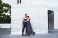 Couple embracing on a rooftop, standing against a white wall, with trees in the background.