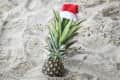 Pineapple with a Santa hat on sandy beach.