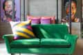 Green velvet sofa with colorful geometric pillows, set against a concrete wall with abstract portraits.