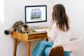 Woman working at a wooden desk with a computer displaying a real estate listing, cat resting beside her.