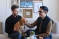 Two people sitting on a sofa with a dog, surrounded by eclectic wall art, books, and a candle on a coffee table.