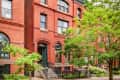 Historic red brick townhouse with arched windows and green doors, surrounded by lush trees and shrubs.