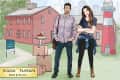 Couple standing with moving boxes in front of a red house for sale, priced at $900,000, with a lighthouse in the background.