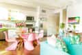 Bright kitchen and living area with pink chairs, glass table, blue sofa, tropical cushions, and neon "Paradise" sign.