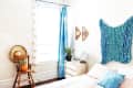 Bedroom with blue curtains, a knitted wall hanging, wooden chair with a potted plant, and a small white dresser.