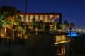 Modern hillside house with large windows, surrounded by palm trees, overlooking city skyline at night.