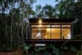 Modern cabin with large windows illuminated from inside, surrounded by dense forest and greenery.
