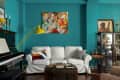 Teal living room with white sofa, colorful pillows, yellow lamp, piano, and eclectic artwork.