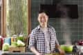 Man in a kitchen holding a tray of asparagus, with a green tiled wall, cookbooks, and vegetables on the counter.
