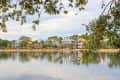 Row of charming houses with palm trees reflected in a calm lake, framed by leafy branches.