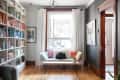Cozy reading nook with a white bench, colorful pillows, a black cat, and bookshelves, under a large window with white curtains.