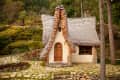 Fairy-tale cottage with a curved shingle roof, wooden door, and stone path surrounded by lush greenery.