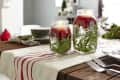 Mason jars with candles, cranberries, and greenery on a wooden table with a striped table runner.