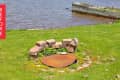 Fire pit with rusted metal cover surrounded by bricks on grassy lakeside area.