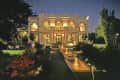 Illuminated mansion at night with arched windows, fountains, and garden path lined with flowers and trees.