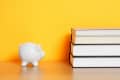 White piggy bank and stack of books on a wooden surface against a yellow background.
