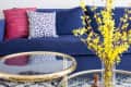 Colorful woven wall hanging above a blue sofa with patterned pillows, glass coffee table, and yellow flowers in a vase.