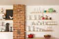Open kitchen shelves with white ceramics, colorful vases, a red pot, and decorative rooster figurines beside a brick column.