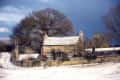 Stone cottage surrounded by snow-covered landscape and bare trees under a cloudy sky.
