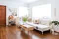 Living room with white sectional sofa, patterned cushions, wooden coffee table, shoe rack, and potted plants on hardwood floor.