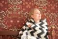 Woman wrapped in a black and white chevron blanket, sitting on a wooden bench against floral wallpaper.