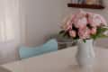 Vase of pink peonies on a white table with a light blue chair in a softly lit room.