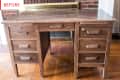 Worn wooden desk with six drawers, placed on a hardwood floor in front of a brick fireplace.