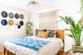 Bedroom with wooden bed, blue throw, macramé wall hanging, hat display, and large potted plant.