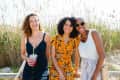 Three women smiling outdoors, one holding a drink, standing in front of tall grass.