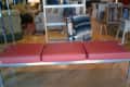 Red cushioned bench with metal frame in a furniture store, surrounded by various chairs and tables.