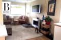 Living room with patterned chairs, wall-mounted TV, fireplace, plant, and dog bed on a beige rug.