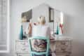 Person with short hair applying makeup at a vintage vanity with a tri-fold mirror and decorative chair.