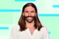 Person with long hair and beard smiling, wearing a white shirt against a pastel geometric background.