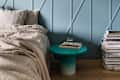 Bedroom with blue paneled wall, beige bedding, turquoise side table, glass of water, books, and stack of magazines.