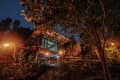 Elevated cabin with large windows surrounded by lush trees and plants, illuminated by warm outdoor lights at dusk.