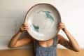 Person in overalls holding a large ceramic plate with green abstract design in front of their face.