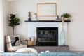 Living room with a white brick fireplace, black mantel, abstract art, potted plants, and a cozy armchair with a basket.