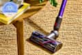 Dyson vacuum cleaner attachment on a woven rug beside a wooden table with books and a potted plant.