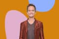 Man in a rust-colored blazer and brown shirt smiling against an abstract orange, pink, and blue background.
