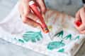 Hand using a Tide stain remover pen on a patterned cloth with green leaves.