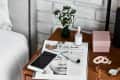 Nightstand with a magazine, smartphone, watch, dropper bottle, pink box, and small vase with flowers.