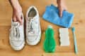 White sneakers on wooden floor with blue cloth, green detergent, sponge, and toothbrush for cleaning.