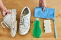 White sneakers with cleaning supplies: blue cloth, green detergent, sponge, and toothbrush on a wooden surface.