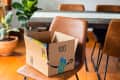 Open Amazon box on a brown chair in a dining room with a wooden table and potted plant.