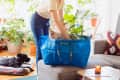 Person placing a plant and pillow into a blue IKEA bag on a sofa, with a dog lying on a rug nearby.