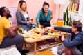 Four friends playing cards around a coffee table with snacks, drinks, and a large jar of cheese balls in a cozy living room.